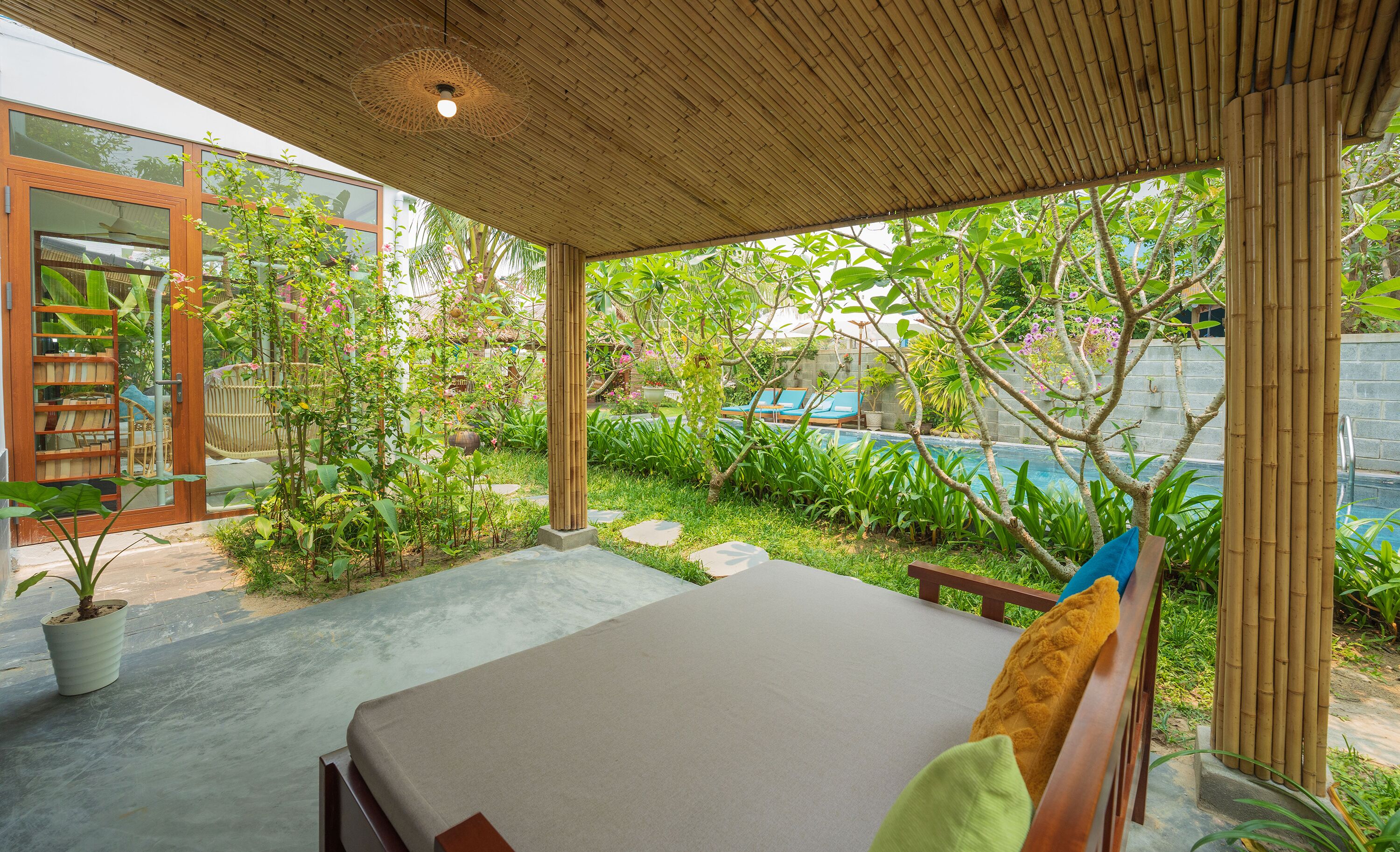 Un espacio de descanso techado al aire libre con una cama, rodeado de exuberante vegetación y vistas a una piscina.