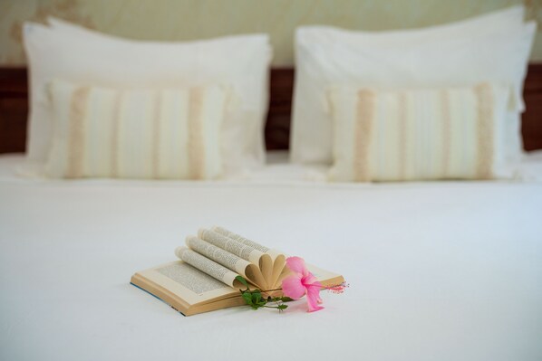 Un lit bien fait, avec une literie blanche et des oreillers à rayures, un livre ouvert décoré d’une fleur rose, et une tête de lit en bois en arrière-plan.