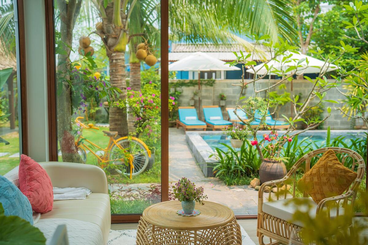 A cozy indoor seating area with a wicker chair and a small round table, a sofa with cushions, and a view of an outdoor swimming pool area with lounge chairs and greenery.