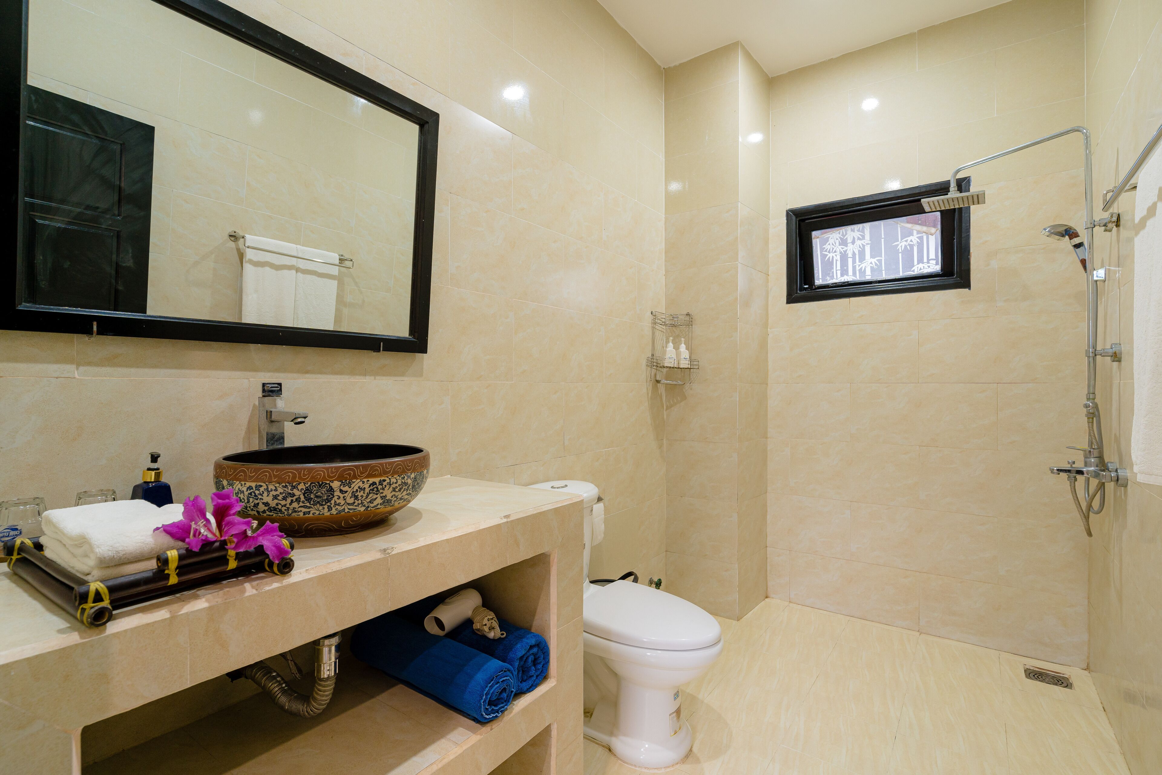 A modern bathroom with a beige tiled wall, a white toilet, a large mirror, a sink with a decorative bowl, and a shower area.