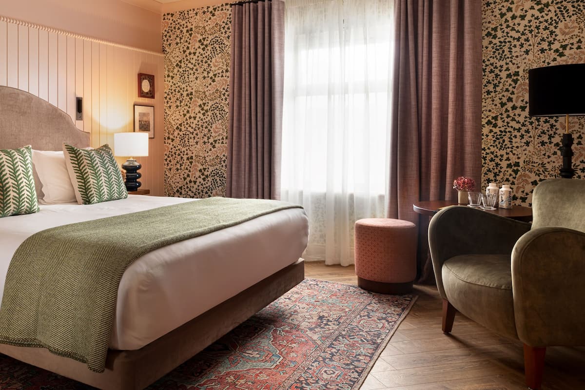 Une chambre d’hôtel avec un grand lit, un pouf rond, une chaise, une table de chevet, une lampe et une fenêtre avec des rideaux.