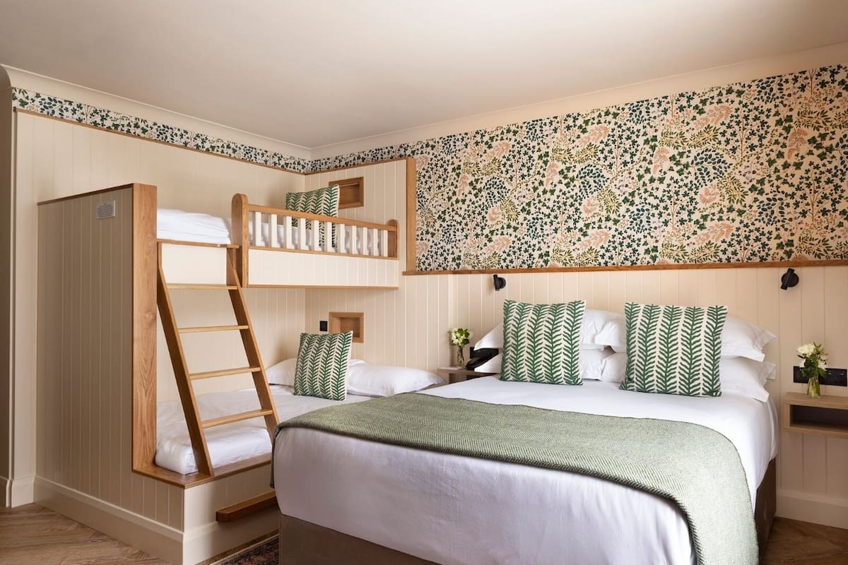 Une chambre avec un lit superposé, un grand lit avec des oreillers assortis, une échelle en bois, un mur à motifs et une petite fenêtre.