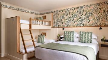 Un dormitorio con una litera, una cama grande con almohadas a juego, una escalera de madera, un papel tapiz con diseño y un ventanal pequeño.