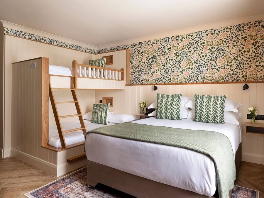 Un dormitorio con una litera, una cama grande con almohadas a juego, una escalera de madera, un papel tapiz con diseño y un ventanal pequeño.