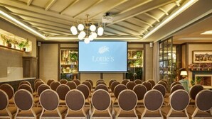 Une salle de conférence avec des rangées de chaises disposées face à un écran où figure le logo de « Lottie’s », un lustre et un espace de réception agrémenté de plantes et d’une cheminée.