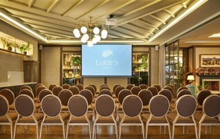 Un salón de conferencias con filas de sillas mirando hacia una pantalla que muestra el logotipo de 'Lottie’s', una araña y una zona de recepción con plantas y una chimenea.