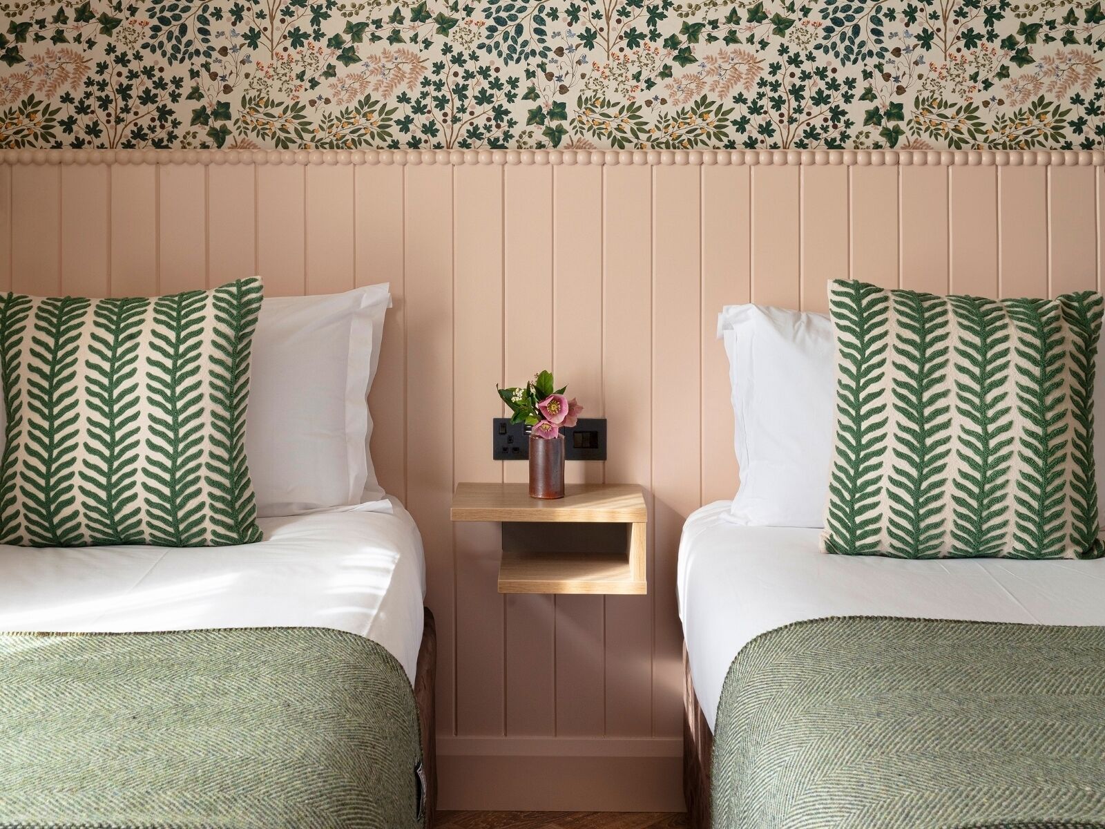 A hotel room with two beds, each with a green leaf-patterned pillow, a small bedside table with a vase of flowers, and a floral wallpaper design.