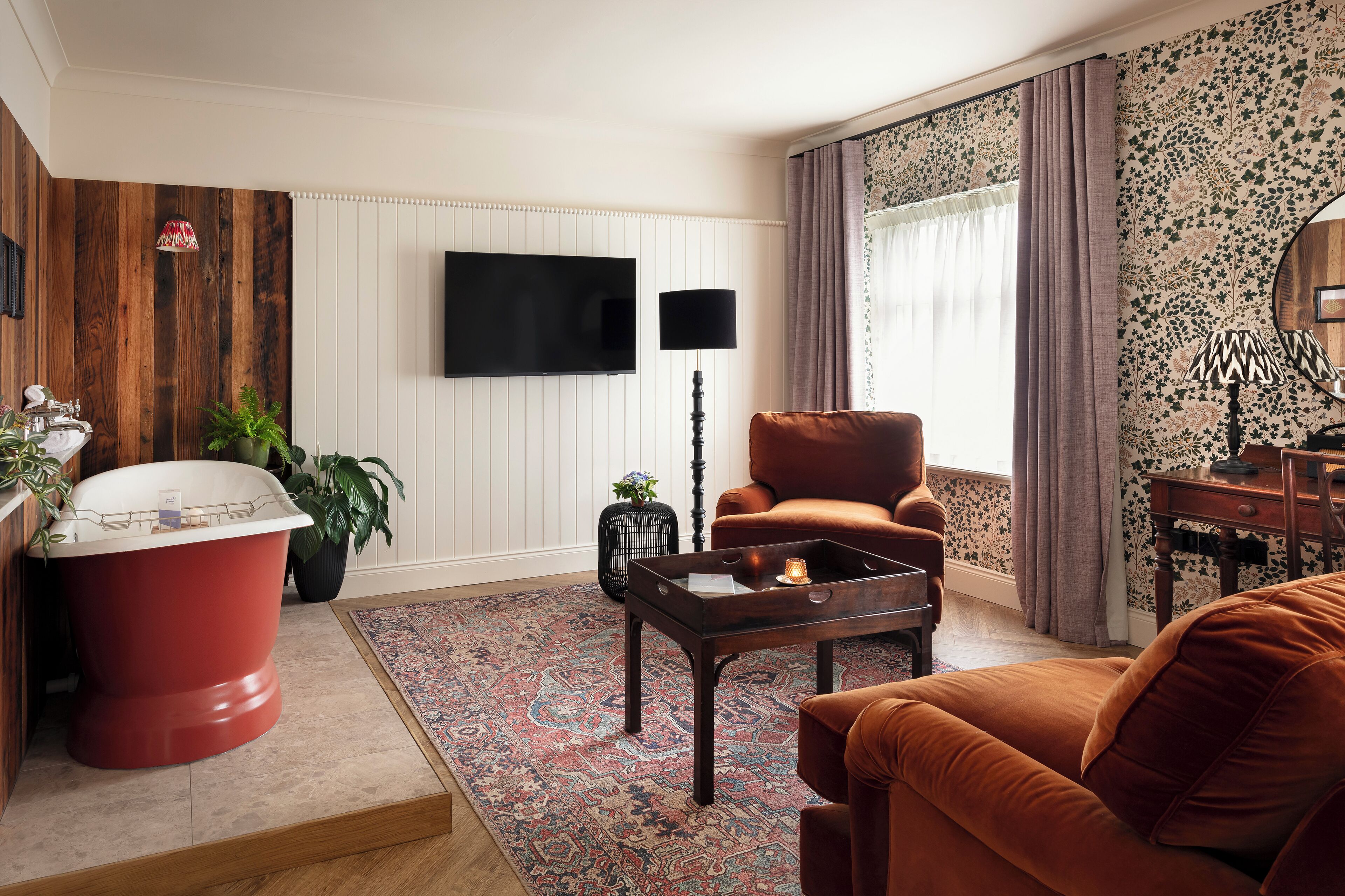 Un salón con una bañera roja, un sillón marrón, una pared de madera, un televisor de pantalla plana y una alfombra estampada.