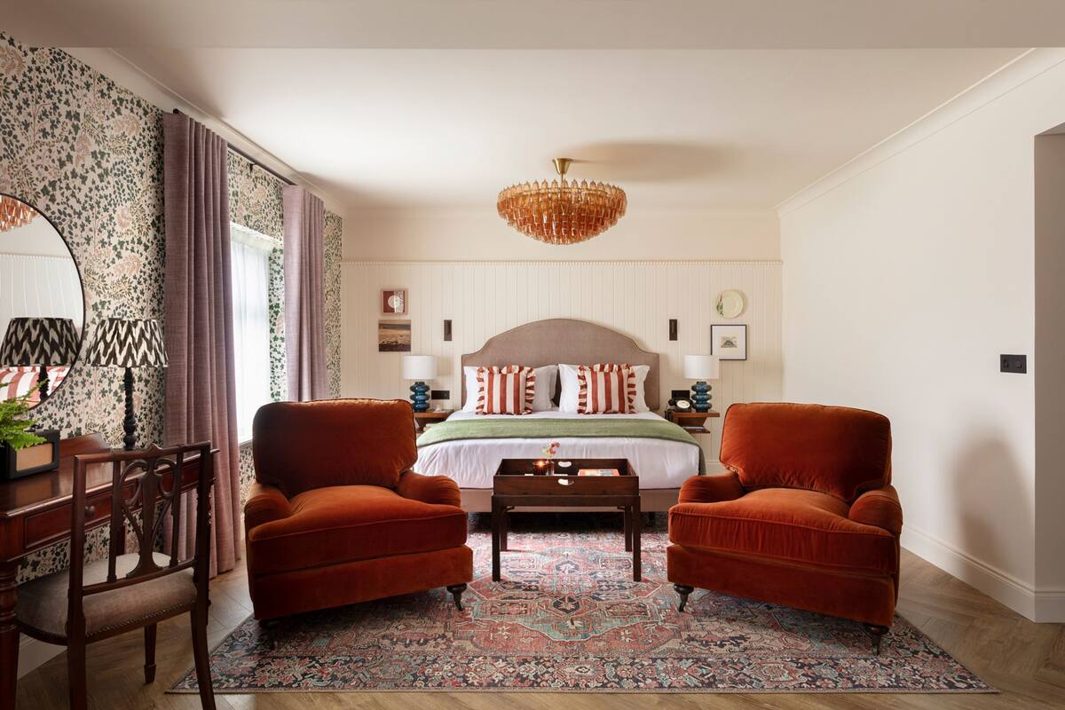 Une chambre à coucher avec un grand lit, deux fauteuils, une table basse en bois, un lustre et un tapis à motifs.