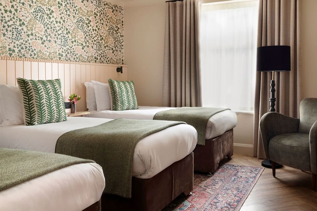 Une chambre d’hôtel avec deux lits, une grande fenêtre avec des rideaux, une lampe sur pied et un tapis à motifs.