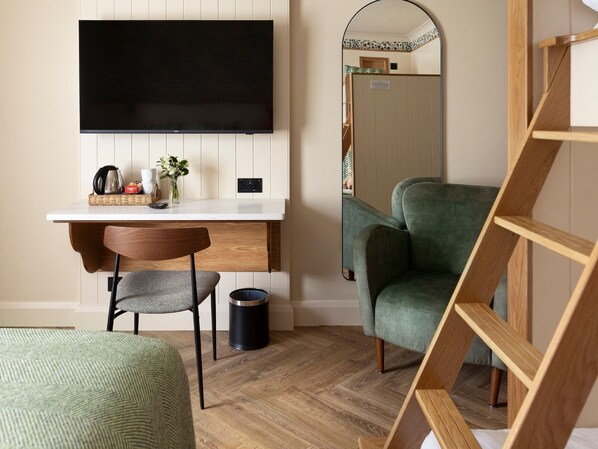 A room with a flat-screen TV mounted on a wall, a wooden desk with a chair, a green armchair, a small trash bin, and a wooden ladder.