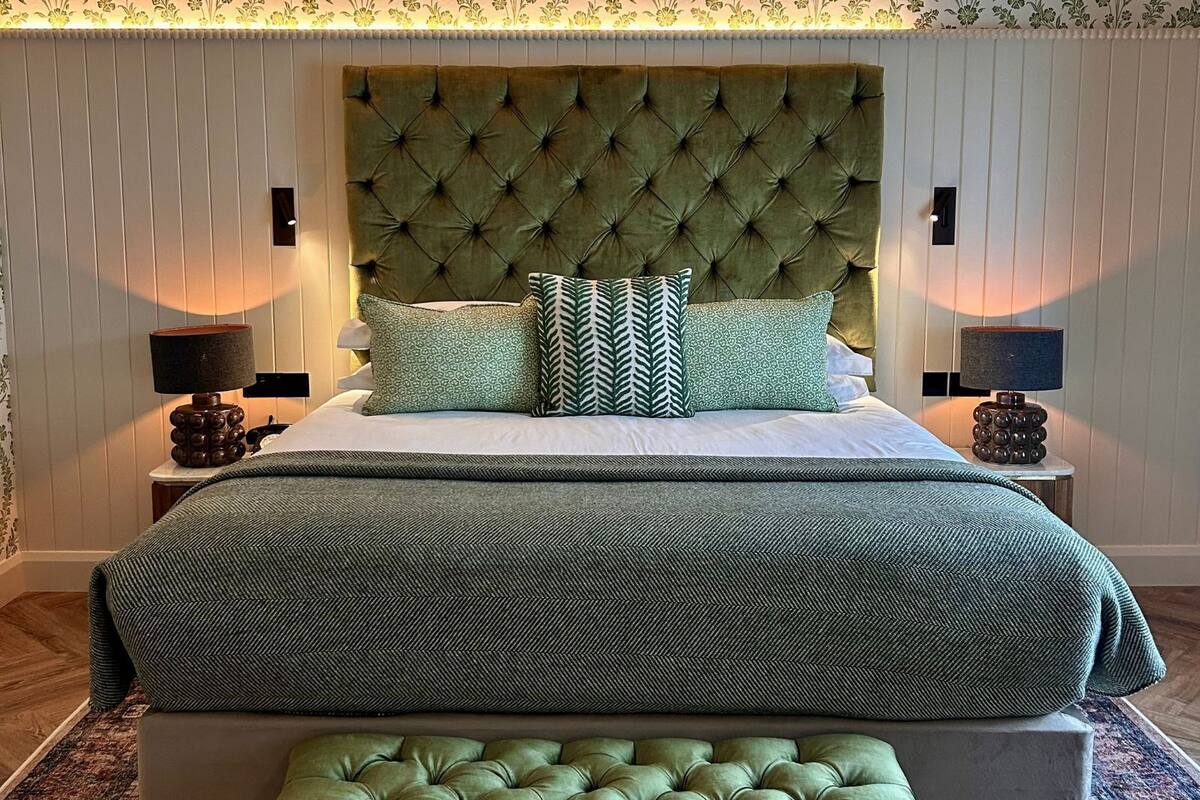 Une chambre à coucher avec une tête de lit capitonnée, un lit avec une literie verte, deux lampes de chevet et un mur à motifs.