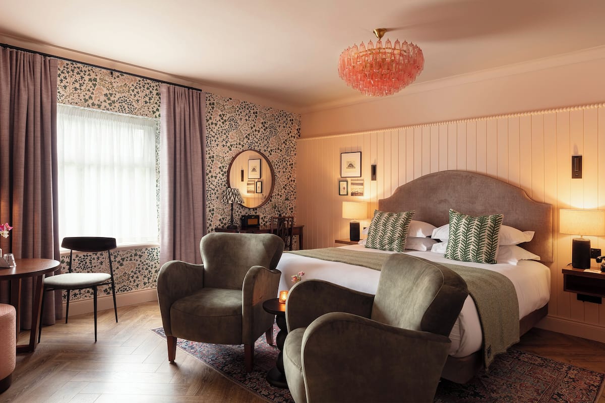 Une chambre d’hôtel avec un grand lit, deux fauteuils, une petite table et un lustre.