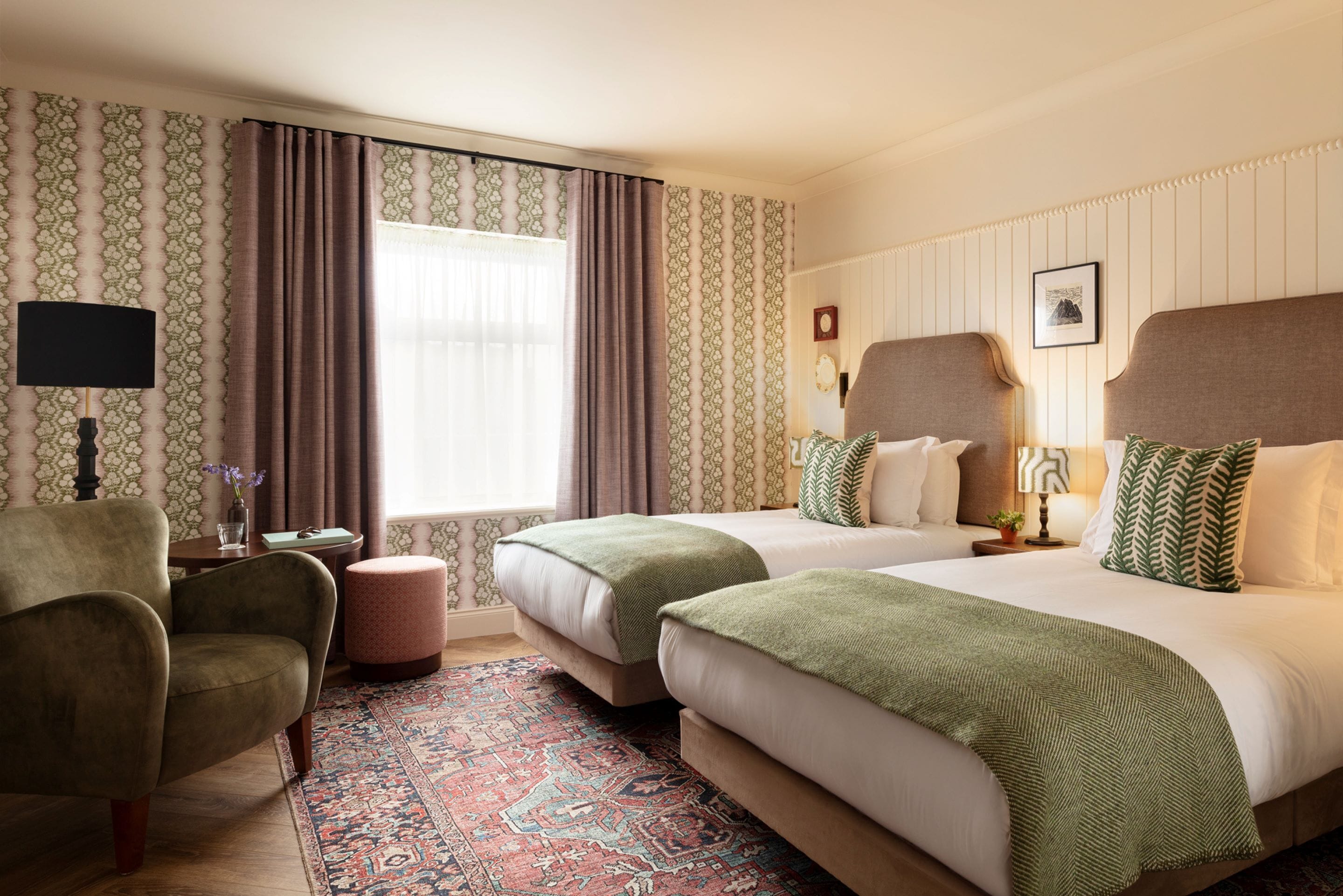 A hotel room with two beds, a chair, a small table, and a patterned rug.