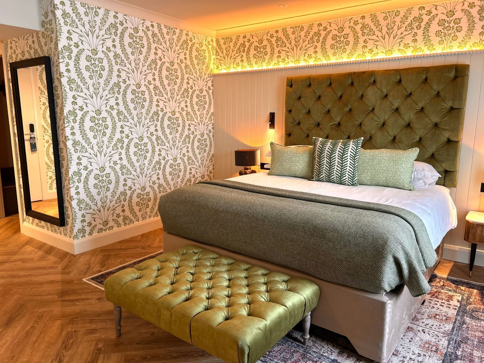Une chambre à coucher avec une tête de lit capitonnée, un grand lit avec de nombreux oreillers, un banc et un mur orné d’un motif.
