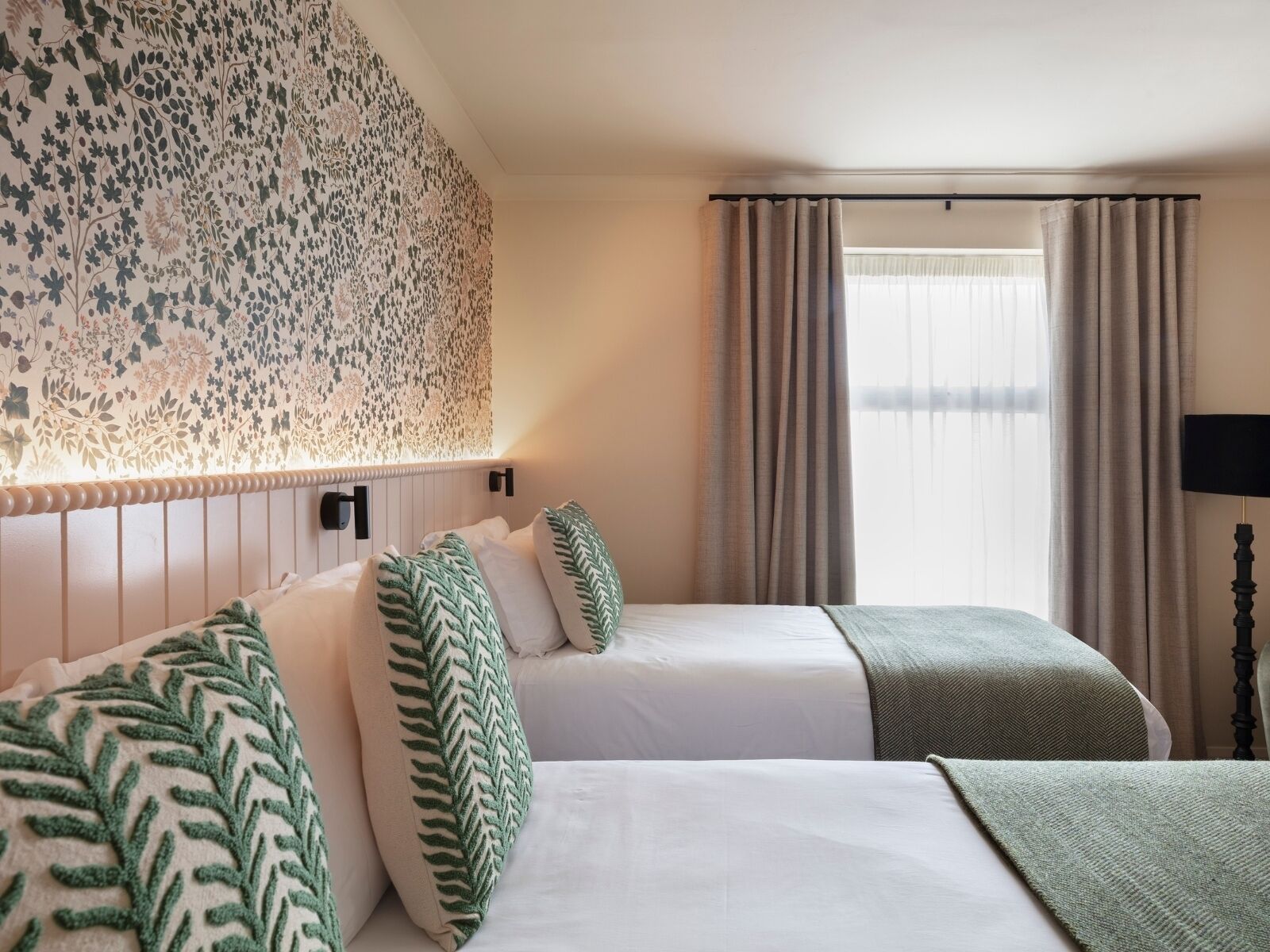 A hotel room with two beds, a floral-patterned headboard, a large window with curtains, and a standing lamp.