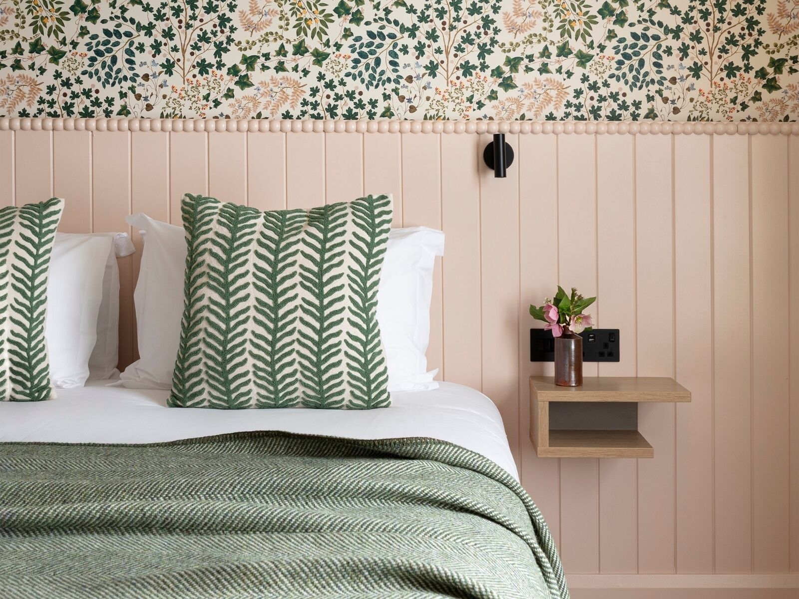 A bedroom with a bed, green patterned pillows, a green herringbone throw, a nightstand with a vase of flowers, and a floral wallpaper.