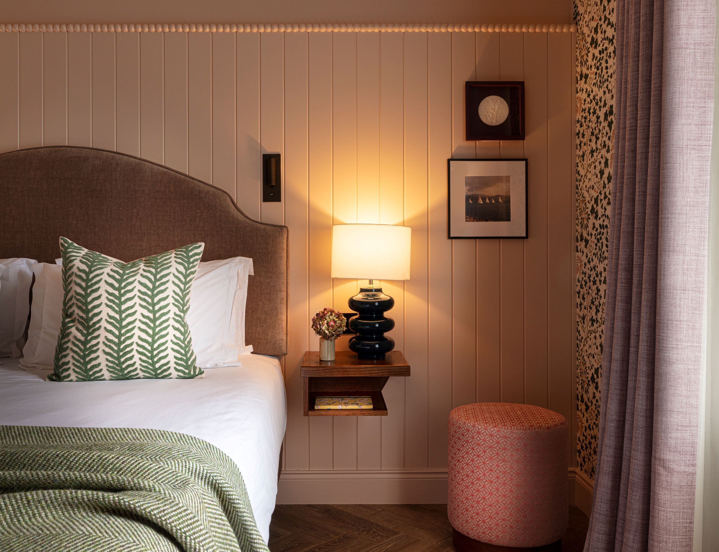 A neatly arranged hotel room with a bed, a bedside table with a lamp, a small stool, and a framed picture on the wall.