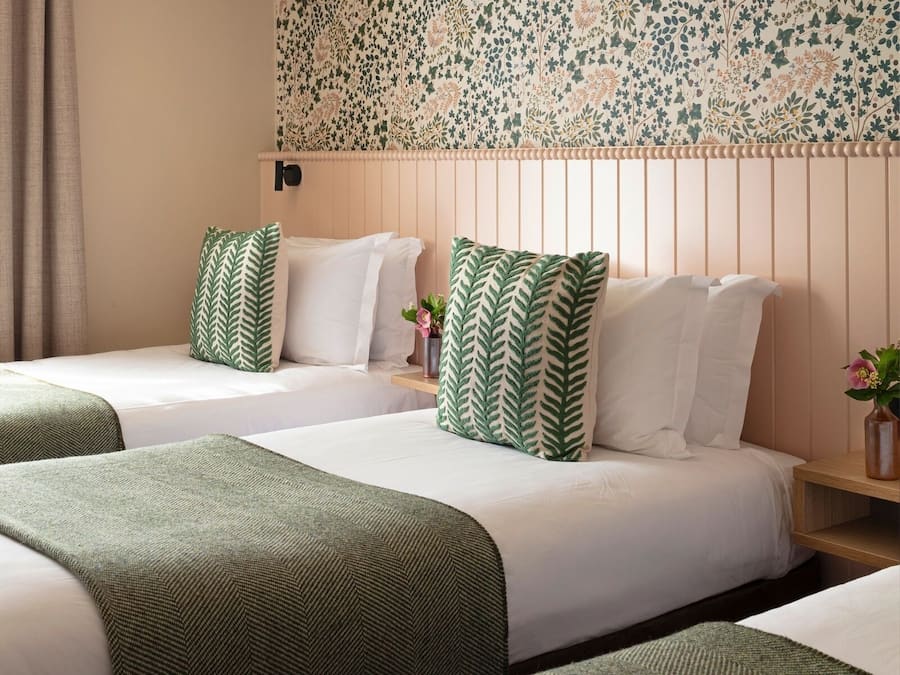A hotel room with two beds, each with green and white patterned pillows, a floral wallpaper, and a wooden headboard.