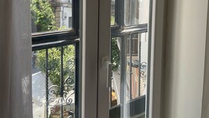 Vista a través de una ventana con barandilla metálica, un balcón y edificios al fondo.
