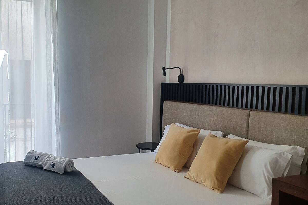 Una habitación de hotel con cama, dos almohadas, mesita de noche, lámpara y ventana con cortinas.
