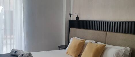 Hotellihuone, jossa on sänky, kaksi tyynyä, yöpöytä, lamppu ja ikkuna, jossa on verhot.