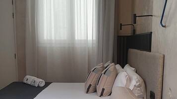 A room with a bed, a sofa, a window with sheer curtains, an air conditioning unit, and a wall-mounted light fixture.