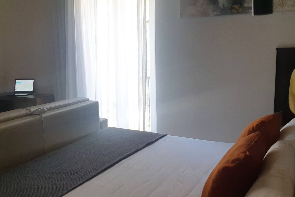Una habitación de hotel con cama, escritorio, computadora portátil, lámpara y ventana con cortinas.