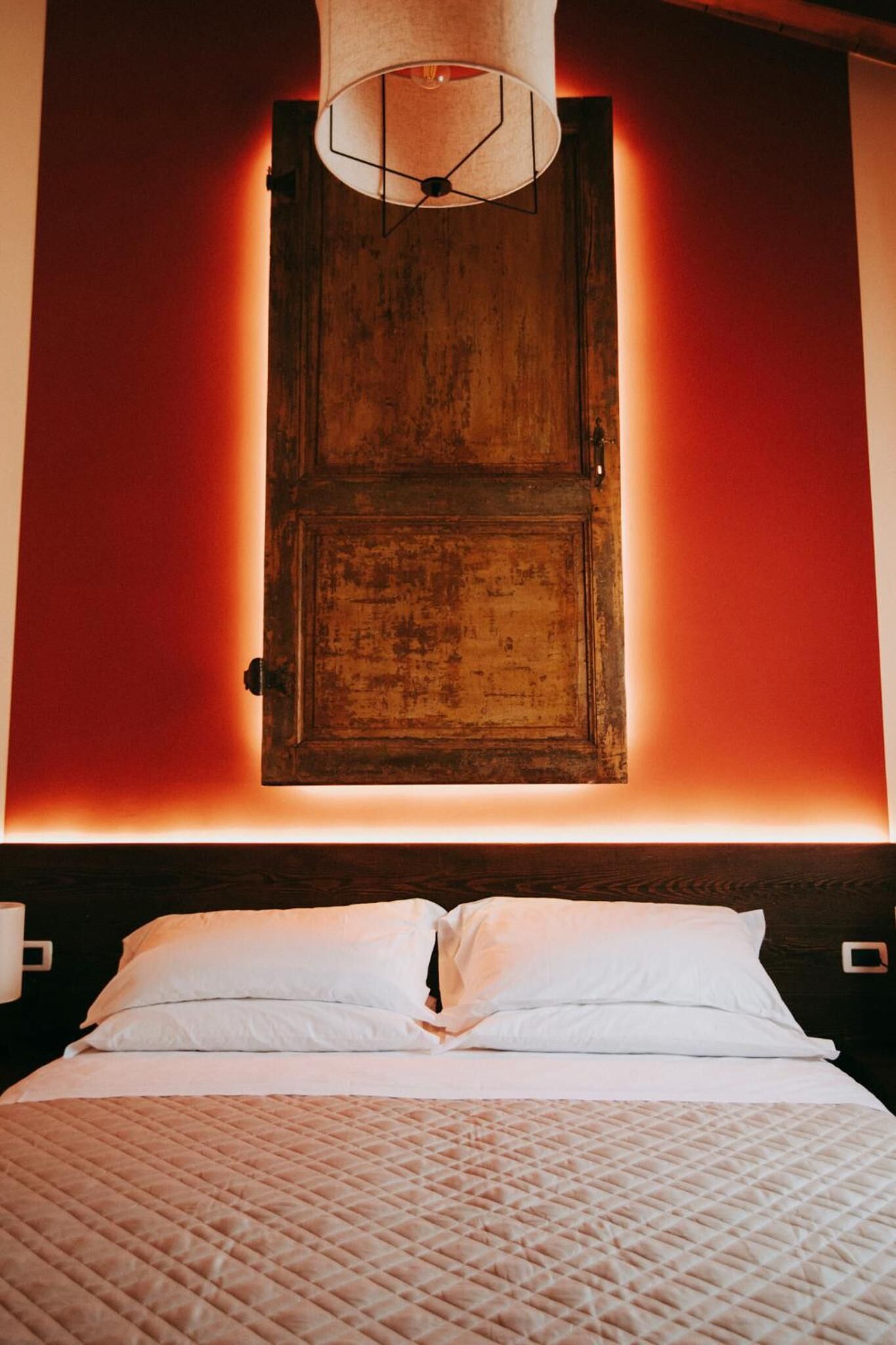 Ein Bett mit weißen Leinentüchern und einer gesteppten Decke, ein Holzschrank mit einer geometrischen Leuchte darüber und eine rote Wand.