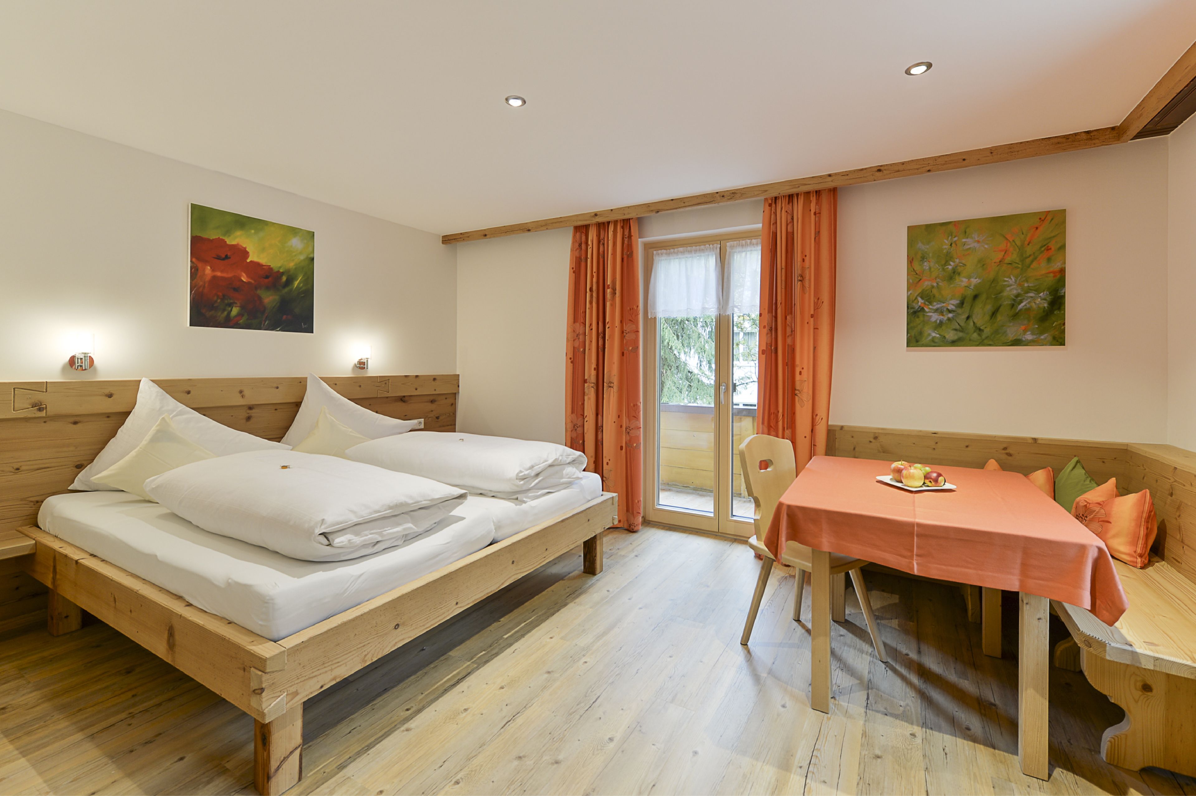 A bedroom with a wooden bed, a table with a tablecloth, and two paintings on the walls.