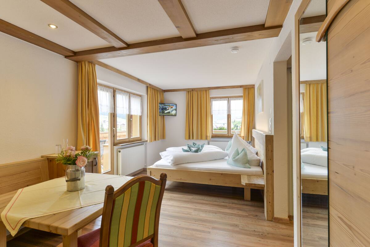 A room with a wooden bed, a table with a vase of flowers, and a television on the wall.