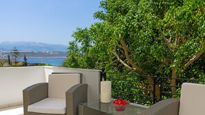 A balcony with wicker furniture and a glass table, overlooking a coastal landscape with trees and mountains. - Villa Plumeria Flower (Chania)