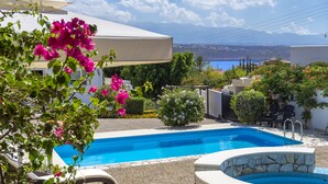 A swimming pool with a view of the sea and mountains, surrounded by greenery and a white building. - Villa Plumeria Flower (Chania)