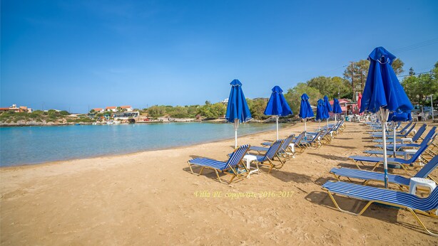 A beach with rows of blue and white striped lounge chairs and matching umbrellas, a sandy shore, and a calm sea. - Villa Plumeria Flower (Chania)