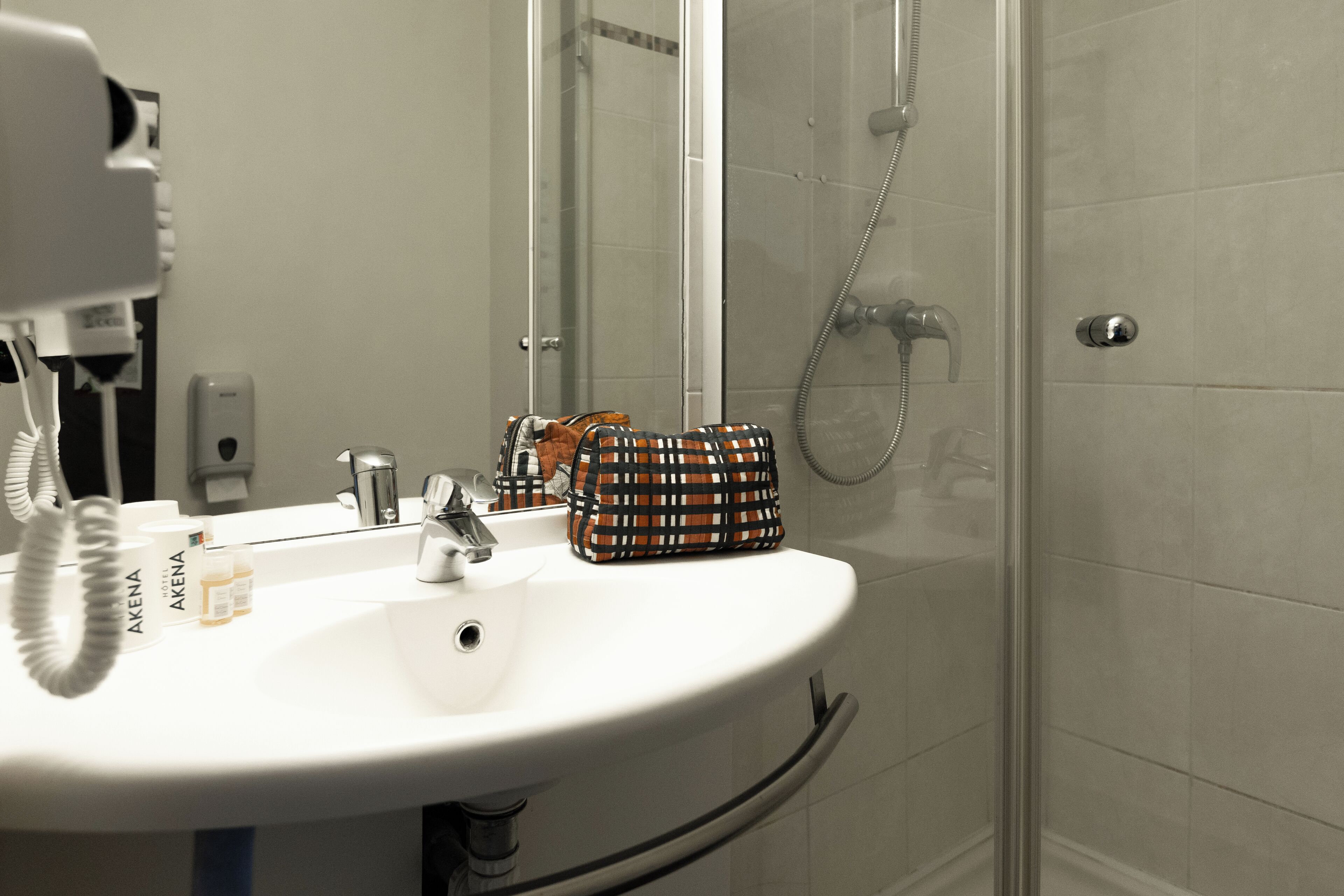 Une salle de bain avec un lavabo blanc, un miroir, une douchette à main et un sac à motifs posé sur le lavabo.