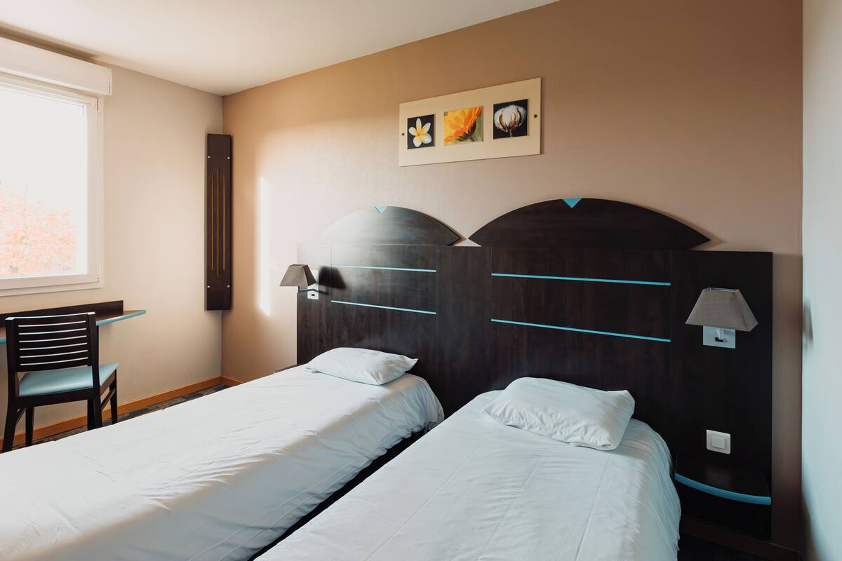 Une chambre d’hôtel avec deux lits simples, un bureau, une chaise et une lampe fixée au mur.