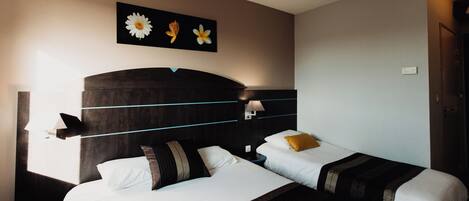 Una habitación de hotel con dos camas, un cabecero oscuro, un cuadro de flores en la pared y una mesita de noche con lámpara.