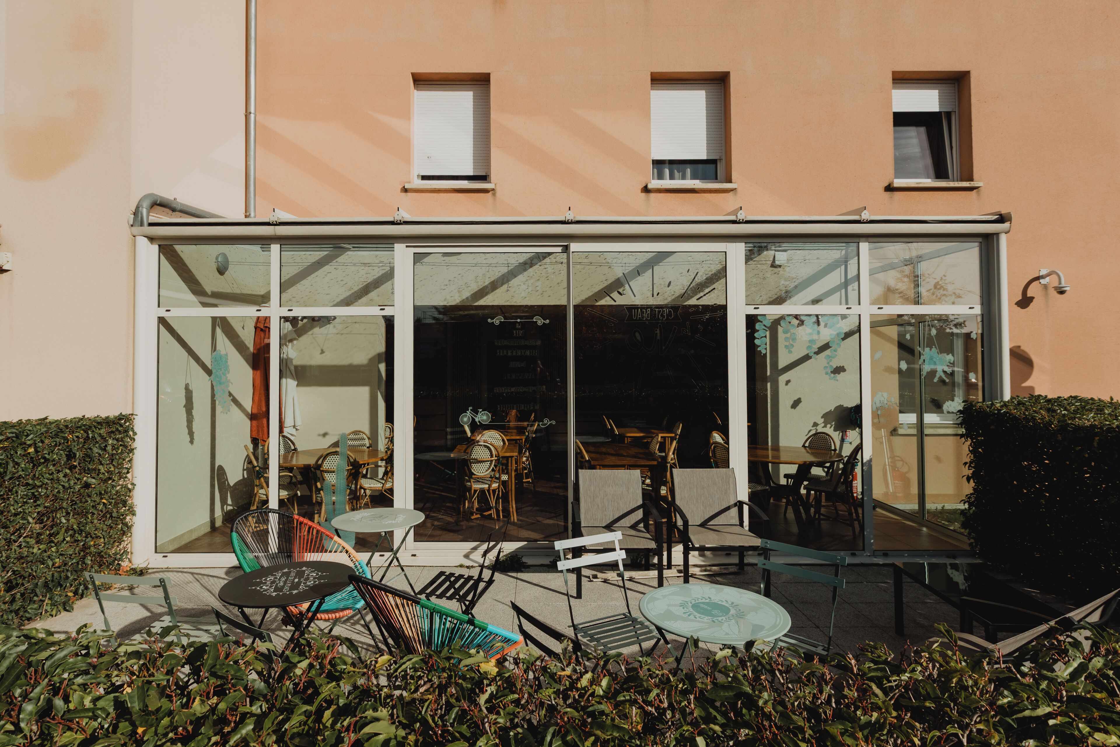 Un edificio con facciata in vetro, area esterna con sedute e tavoli, e un piccolo giardino.