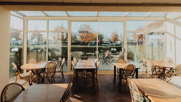 Un patio soleado con mesas y sillas, grandes puertas de cristal y vistas a la calle y árboles.