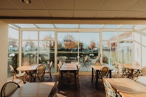 Een zonnig terras met tafels en stoelen, grote glazen deuren en uitzicht op de straat en bomen.