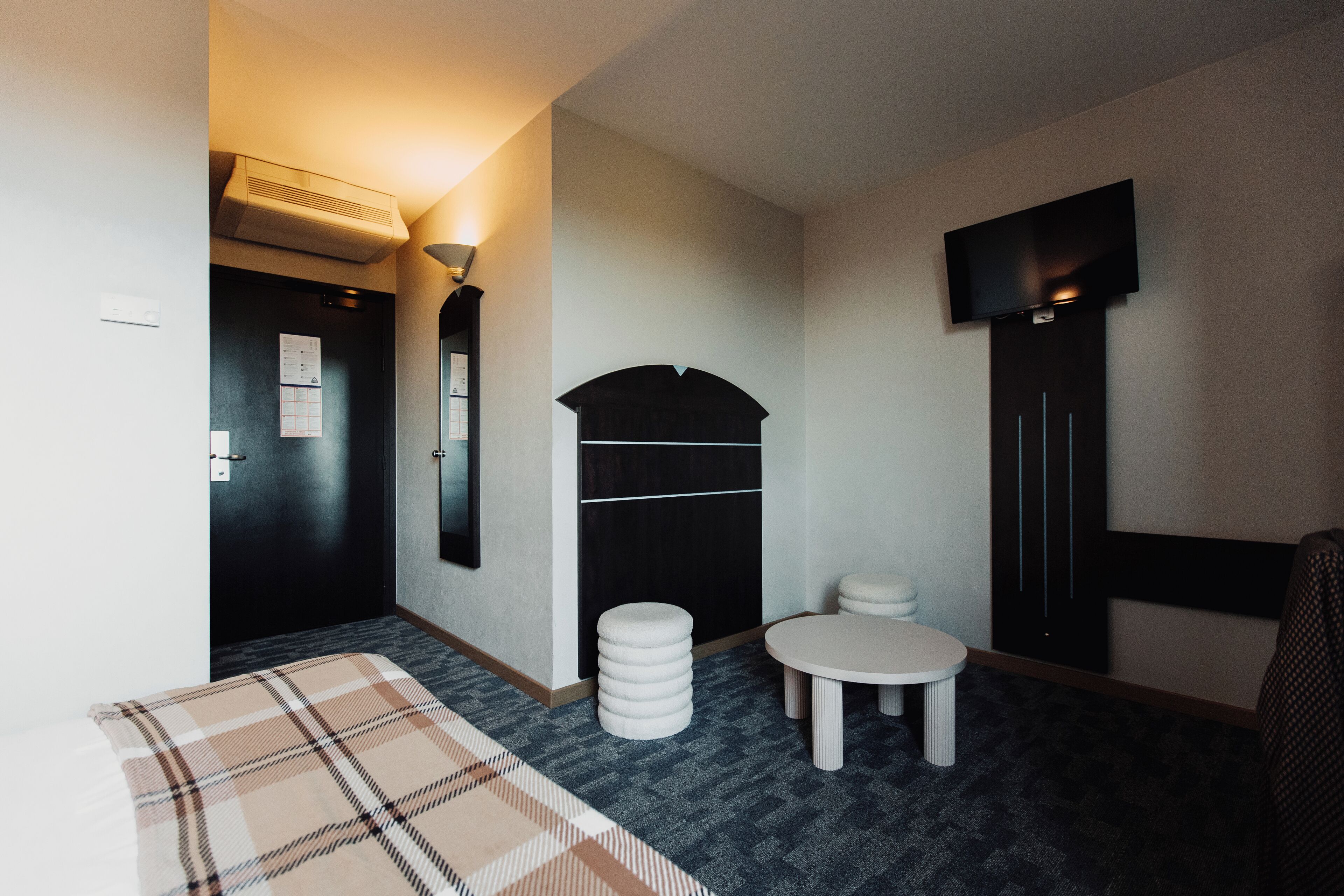 A hotel room with a bed, a small round table, a sofa, a TV mounted on the wall, and a door with a calendar on the wall.