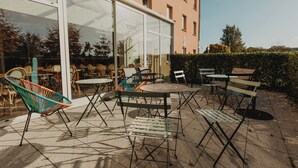 Un patio al aire libre con mesas y sillas, una entrada con puerta de cristal y un edificio de fachada color crema.
