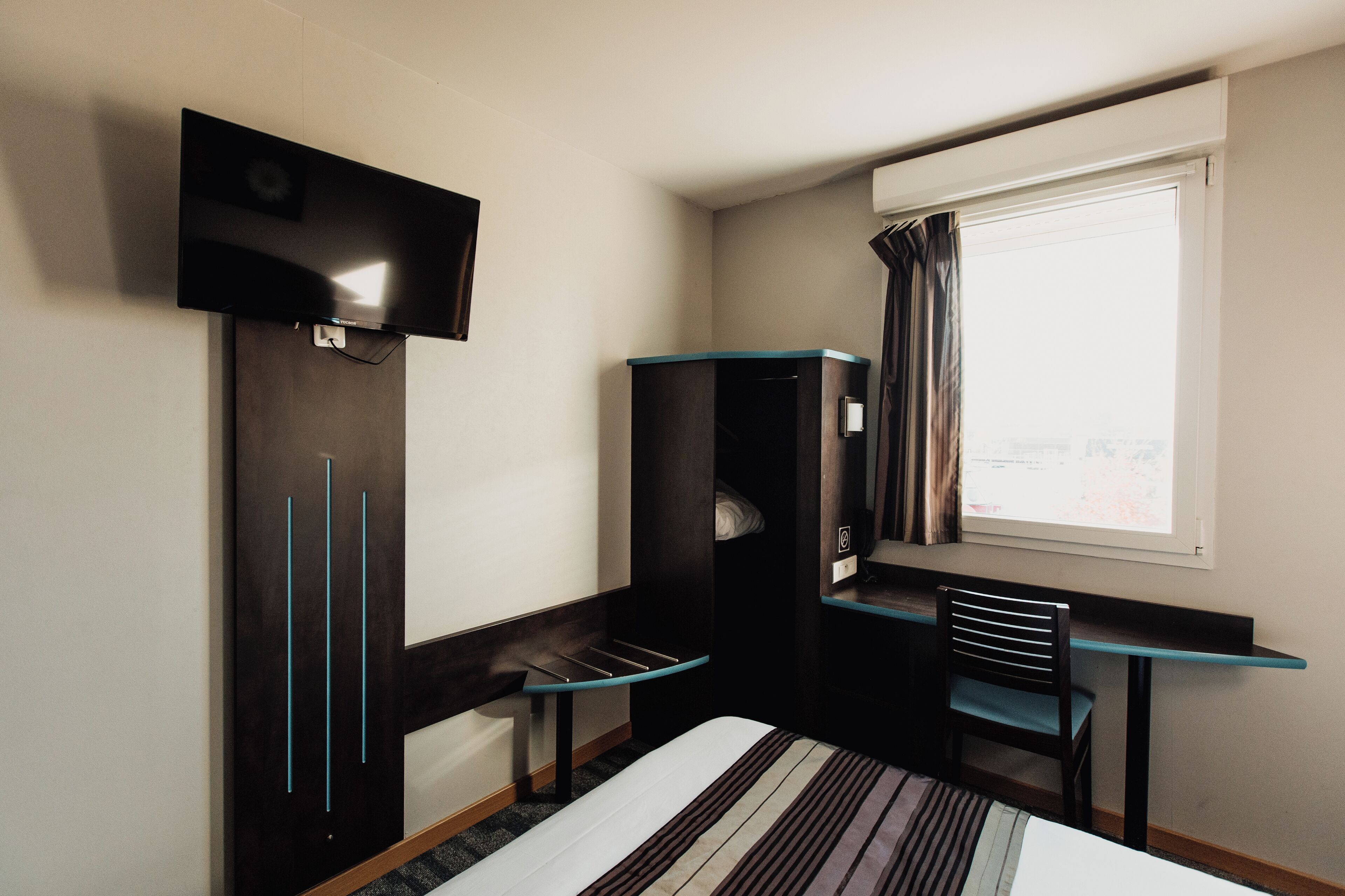 Ein kleines Hotelzimmer mit Bett, Schreibtisch, Stuhl, Fernseher und einem Fenster mit Vorhängen.