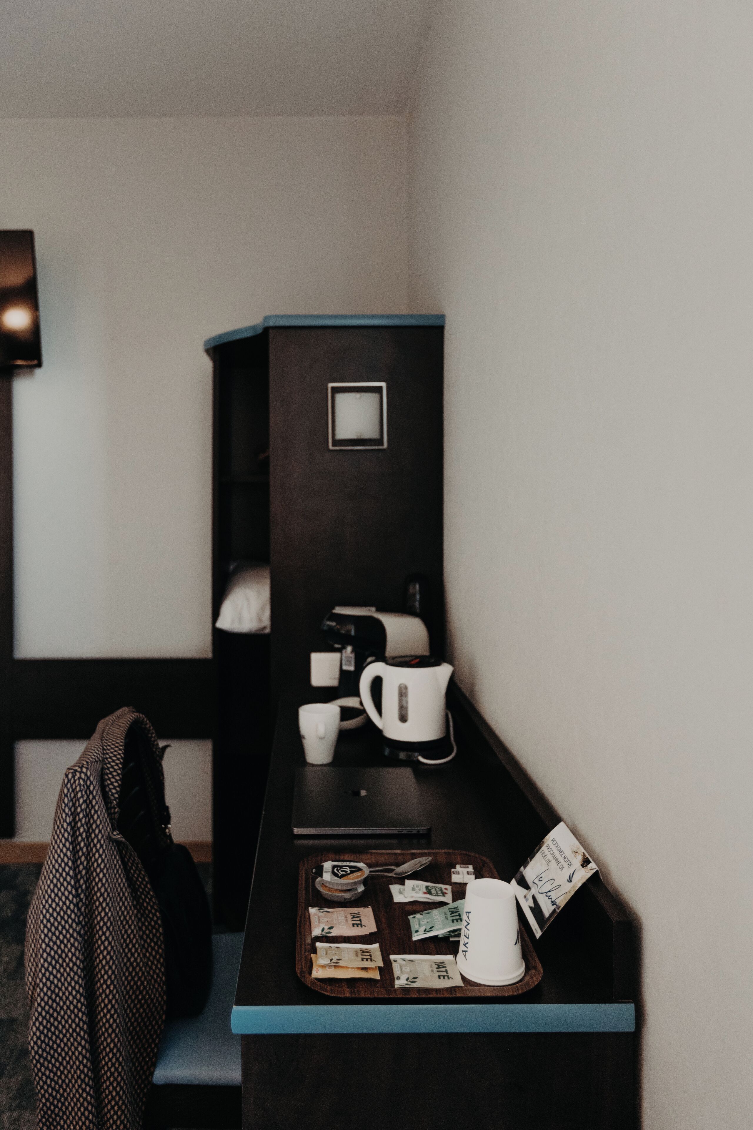 A hotel room with a dark wooden desk, a coffee maker, a tray with various items, and a bag hanging on the wall.