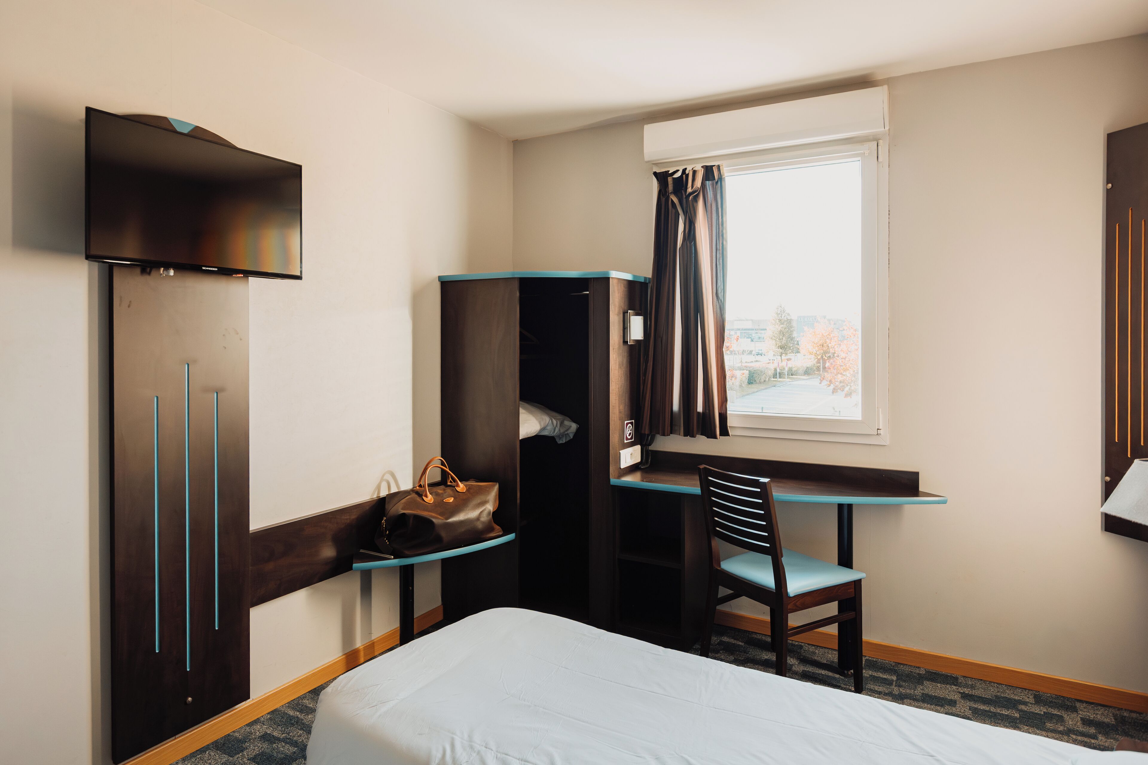 A hotel room with a bed, a desk with a chair, a wardrobe, a TV, and a window with curtains.