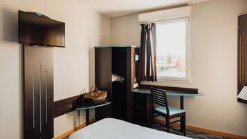A hotel room with a bed, a desk with a chair, a wardrobe, a TV, and a window with curtains.