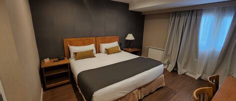 A hotel room with a large bed, wooden headboard, two bedside tables, a lamp, a radiator, and a window with curtains.