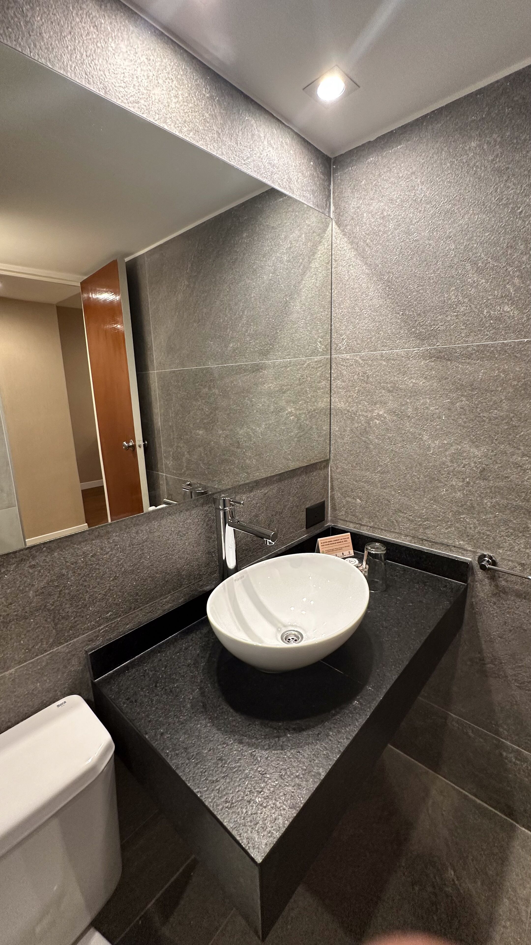 Un baño moderno con un lavamanos blanco, encimeras oscuras y un espejo grande.