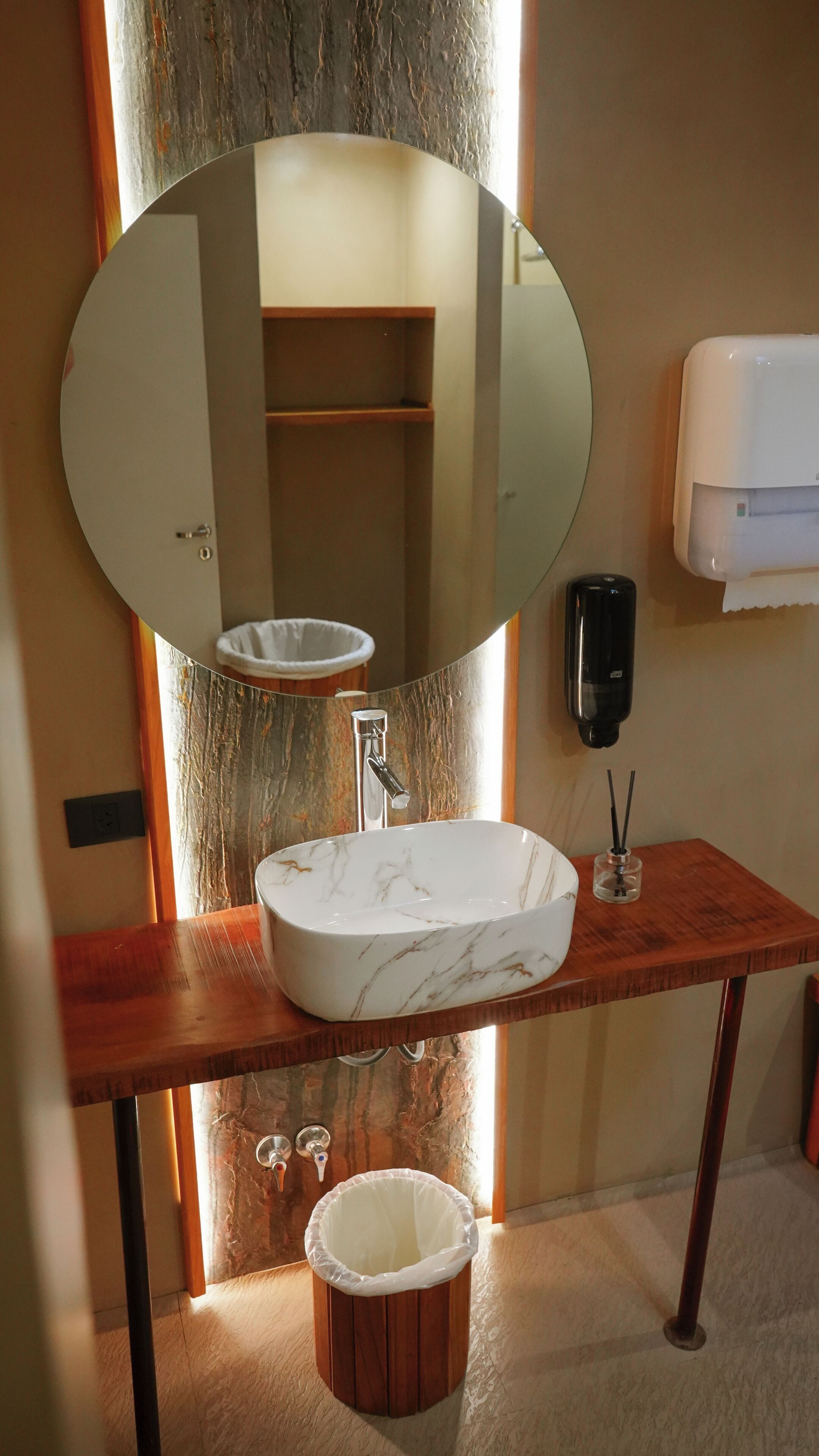 A bathroom with a marble sink, a wooden vanity, a large mirror, a trash bin, and a hand sanitizer dispenser.