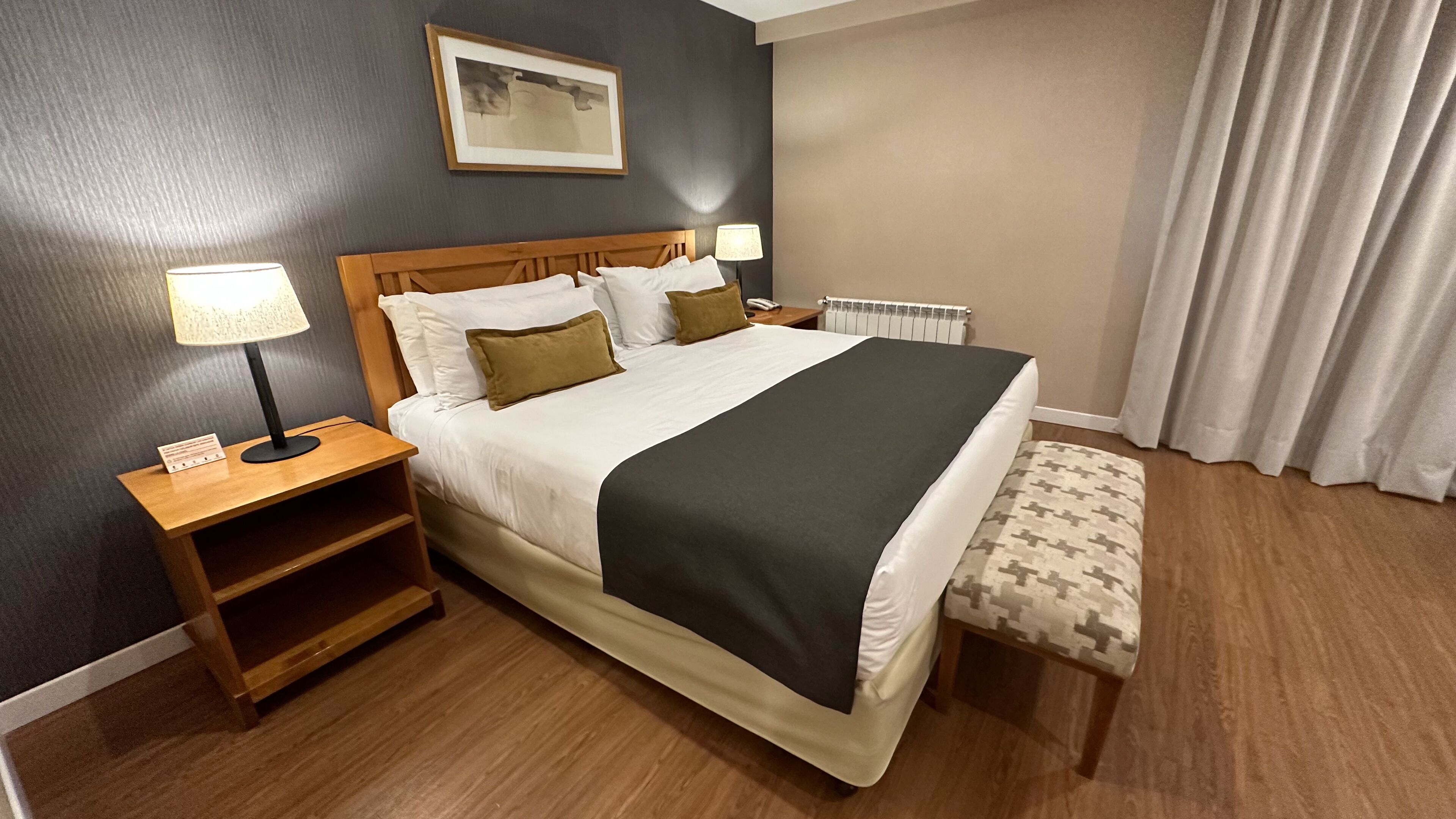 Una camera d'albergo con un letto grande, due comodini, una lampada da comodino, un termosifone e una finestra con tende.